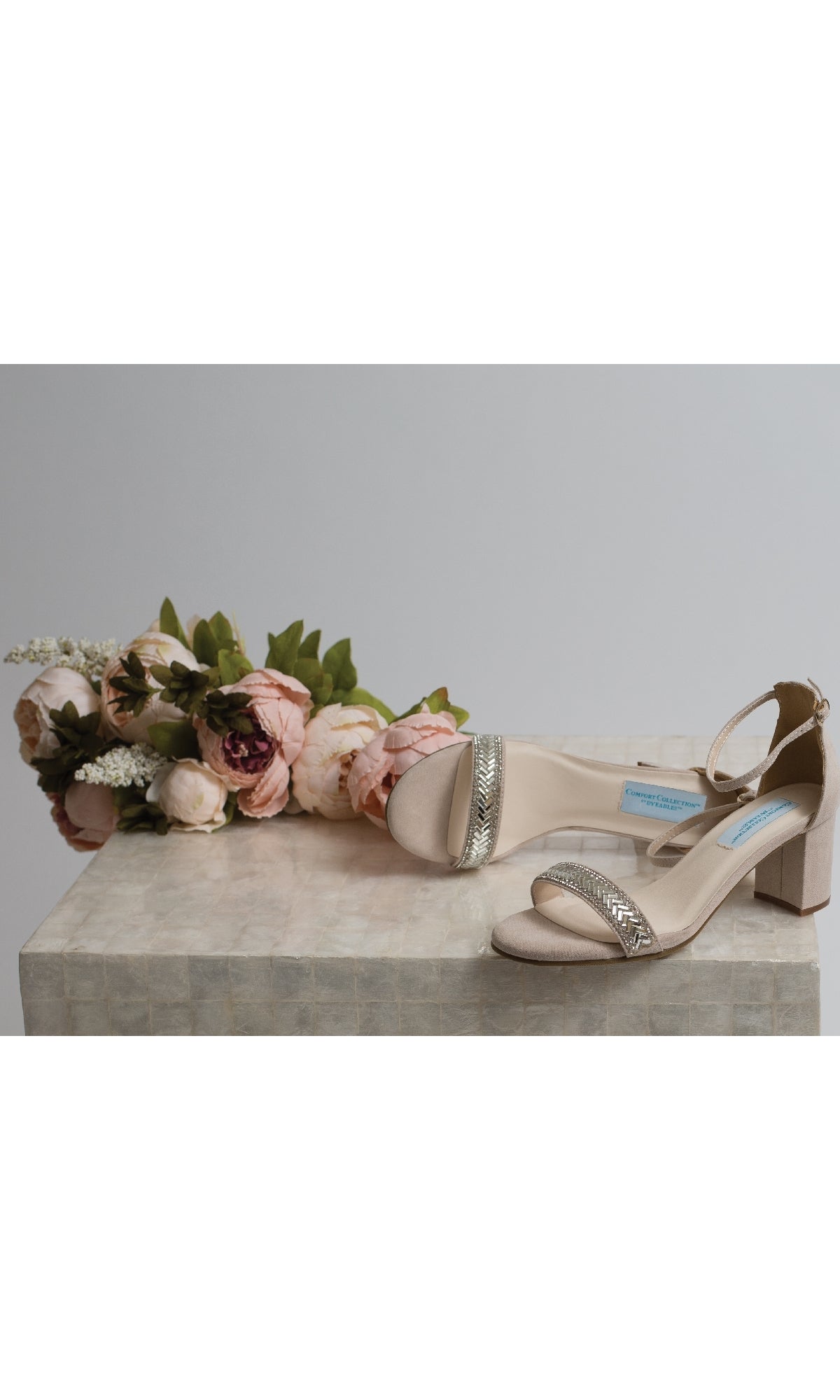 The January Beige Prom Shoes: Dyeables 4368 feature an elegant ankle strap and open-toe block heel, displayed on a light surface beside a bouquet of artificial pink flowers.